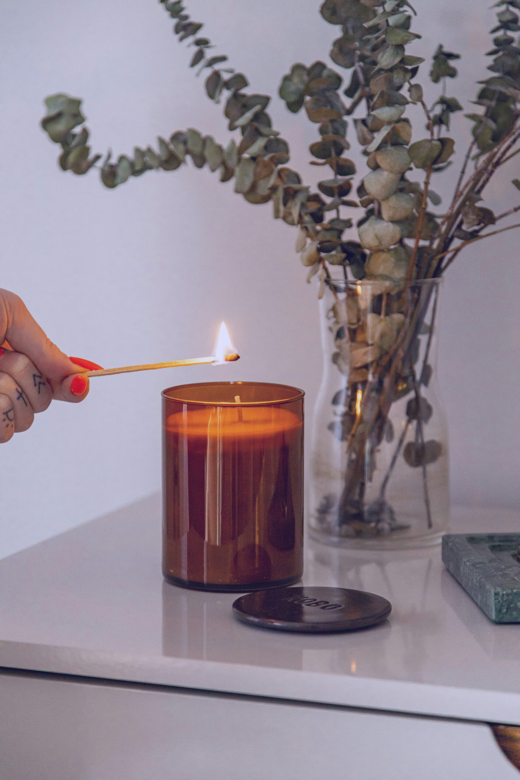 Hand using a long match to light a candle sitting on a table next to a vase of dried eucalyptus