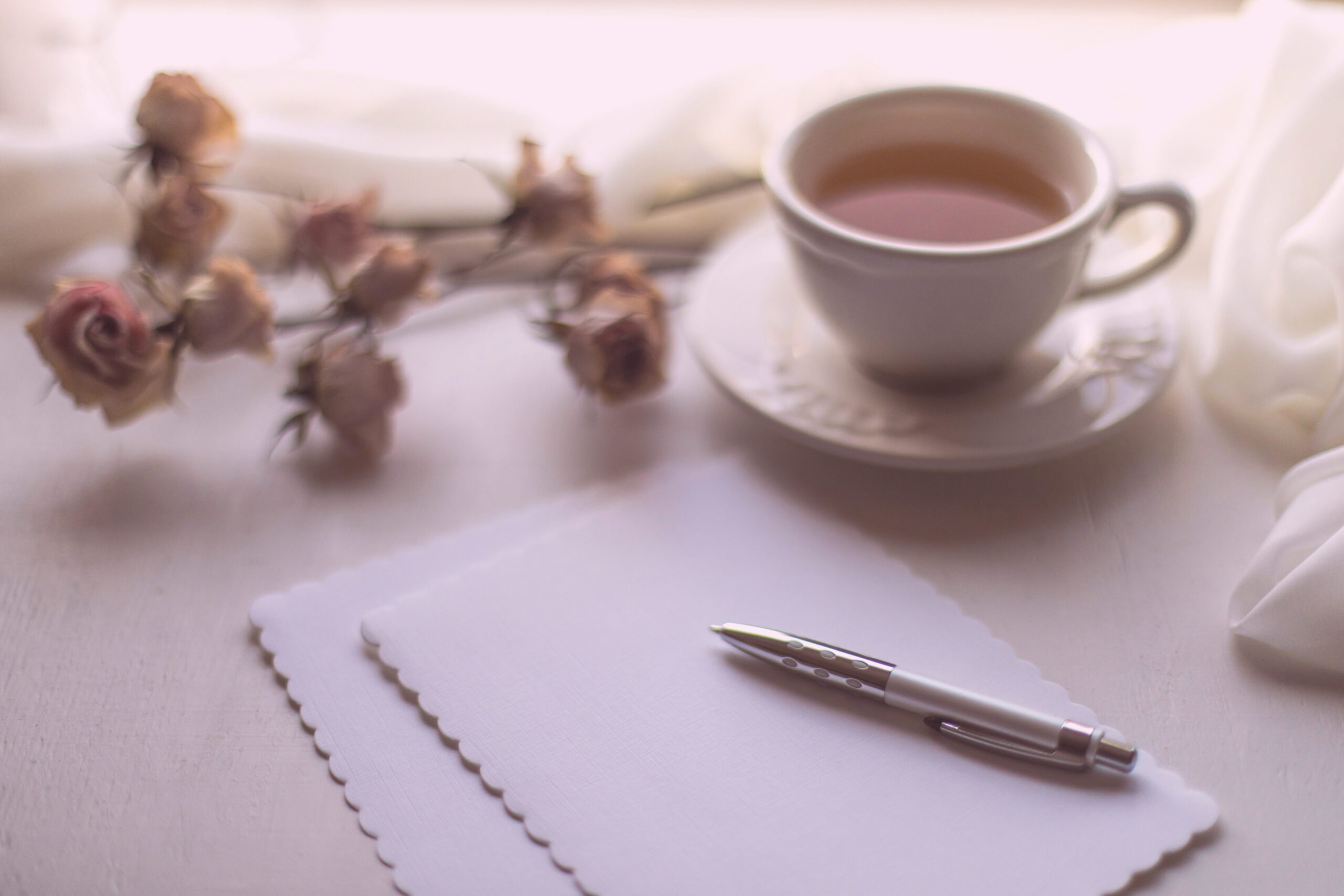 Note paper and a pen sitting on a white desk with a cup of tea and bundle of dried flowers